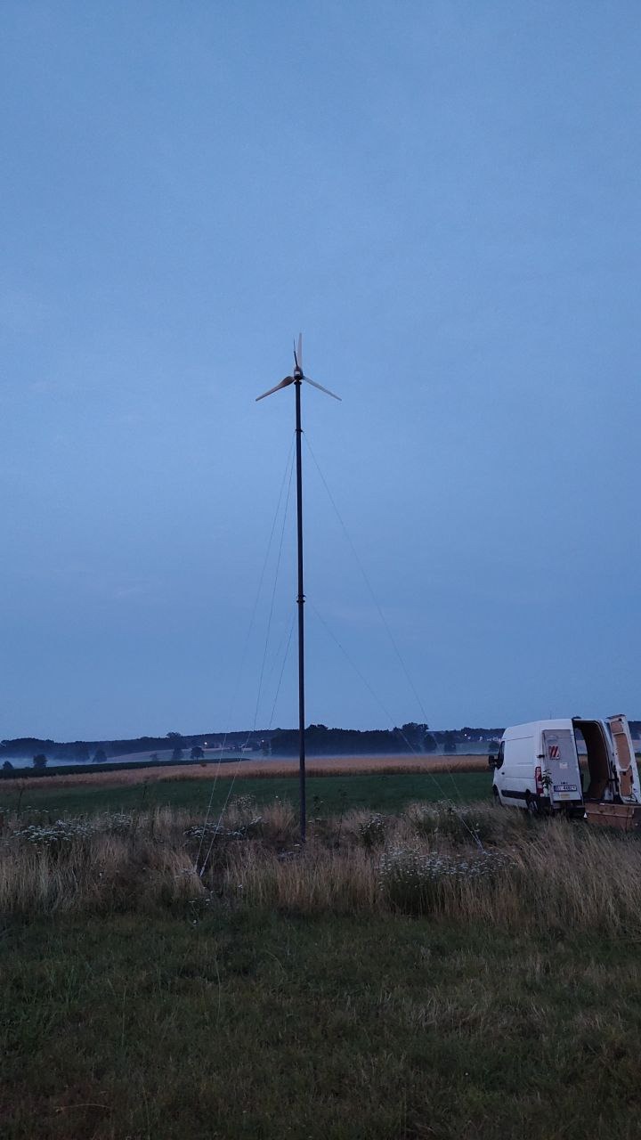 Niska turbina wiatrowa na łące o zmierzchu, z białym vanem serwisowym z otwartymi drzwiami w tle, sugerującym prace instalacyjne lub konserwacyjne.