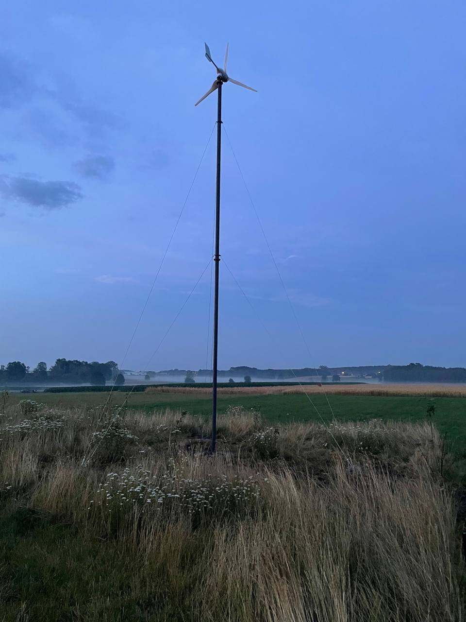 Samodzielna turbina wiatrowa na łące o zmierzchu, wsparta na linach, z widoczną mgłą w oddali, oświetlona delikatnym światłem.