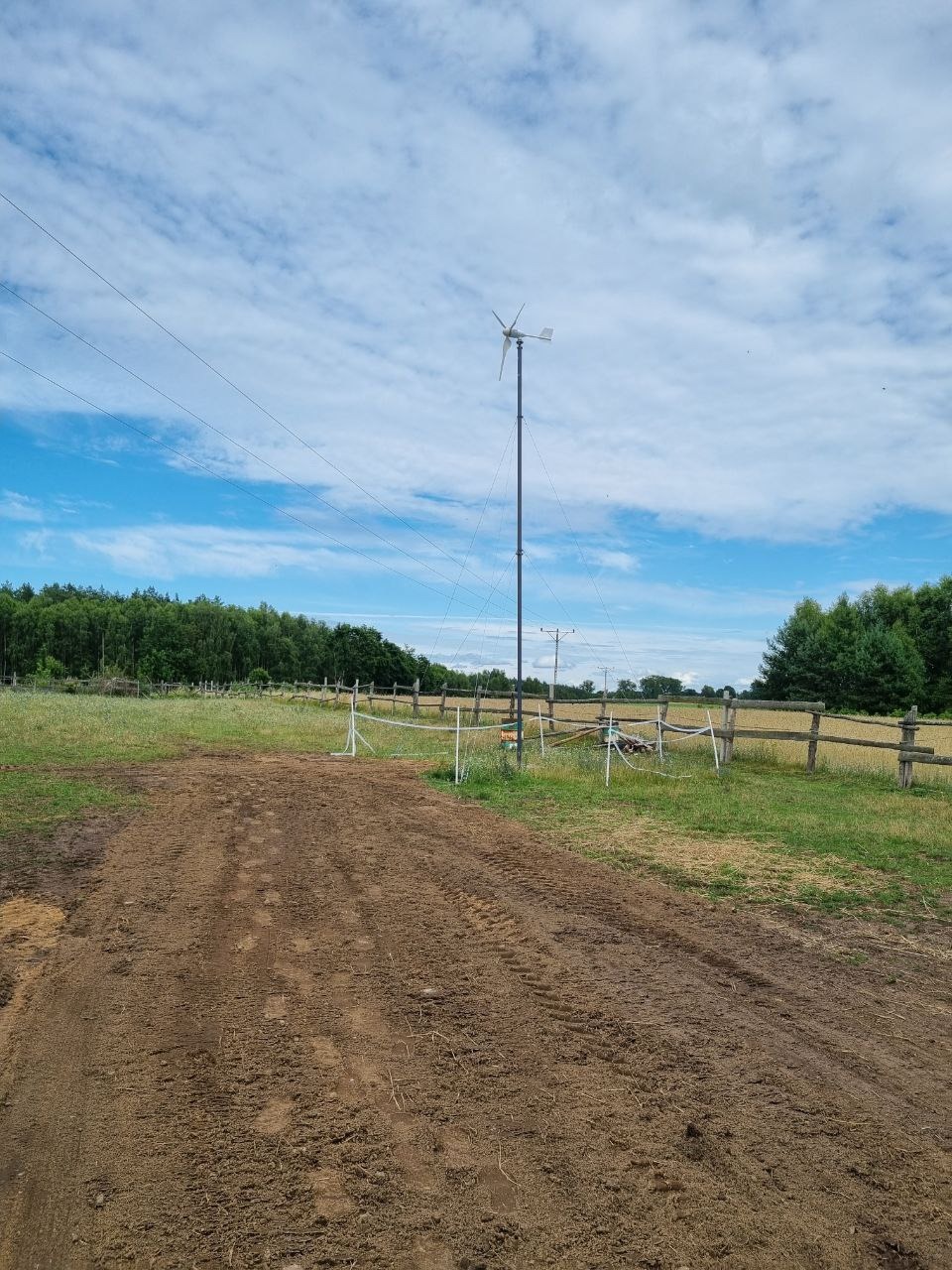 Niska turbina wiatrowa na łące, widok z perspektywy ziemi, z drewnianym ogrodzeniem w tle i błękitnym niebem.