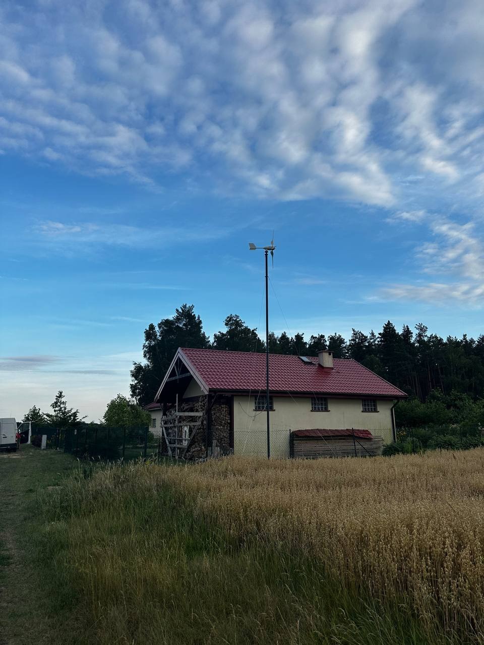 Dom z czerwoną dachówką i przydomową turbiną wiatrową na tle błękitnego nieba z chmurami, otoczony polami i lasem. Energia odnawialna w krajobrazie.