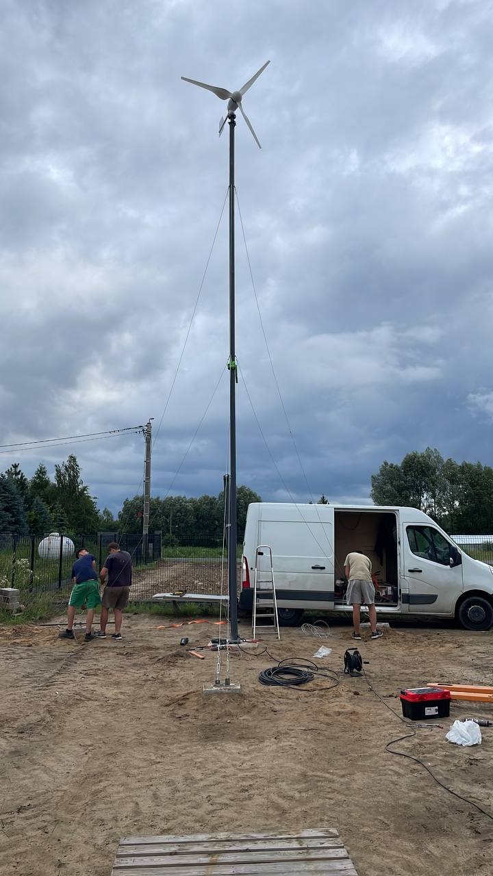 Montaż turbiny wiatrowej: ekipa przy pracy, wysoki maszt z turbiną na tle pochmurnego nieba, bus serwisowy z otwartymi drzwiami i narzędzia na ziemi.