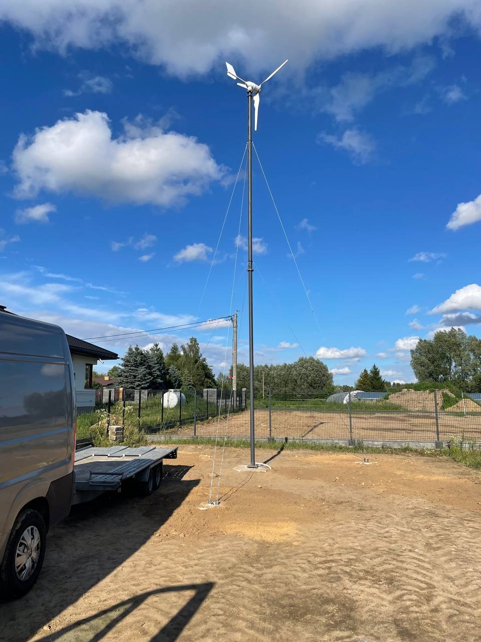 Montaż małej turbiny wiatrowej na posesji, widoczny transport na przyczepie i częściowe ujęcie domu. Błękitne niebo z chmurami w tle.