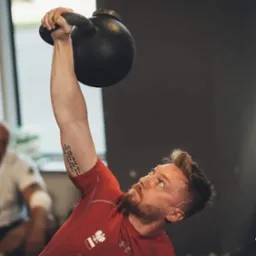 Mężczyzna w czerwonej koszulce unosi kettlebell nad głową, prezentując siłę i determinację. W tle drugi mężczyzna obserwuje trening.