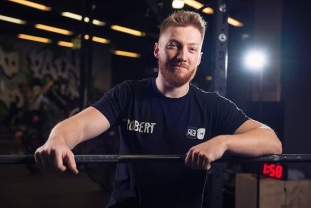 Mężczyzna z brodą w czarnej koszulce opiera się o sztangę w siłowni, w tle graffitti na ścianie i oświetlenie sufitowe. Widoczny zegar z czasem 1:58.