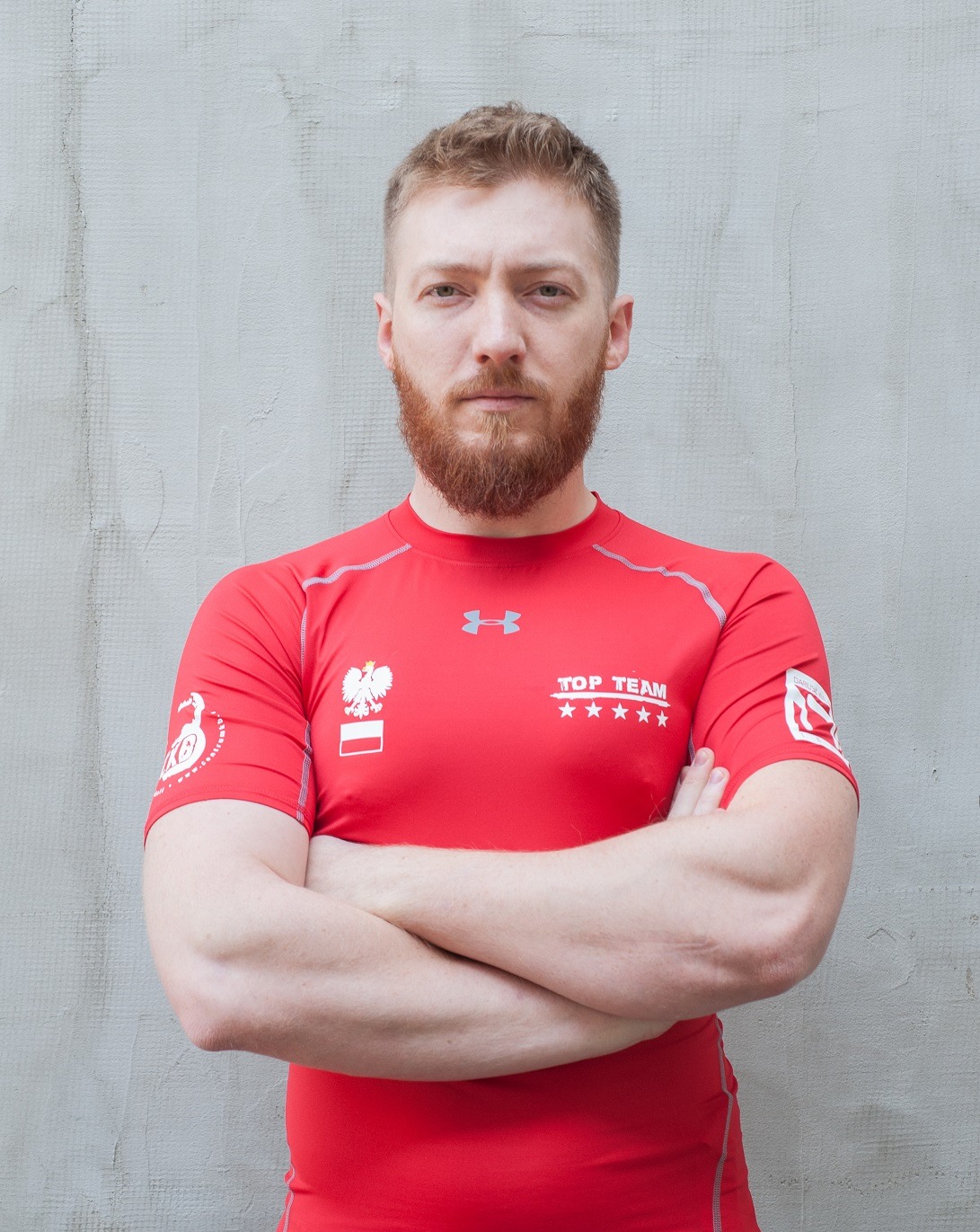 Mężczyzna w czerwonej koszulce sportowej z godłem Polski i napisem 'TOP TEAM' na tle szarej ściany, skrzyżowane ręce, broda.