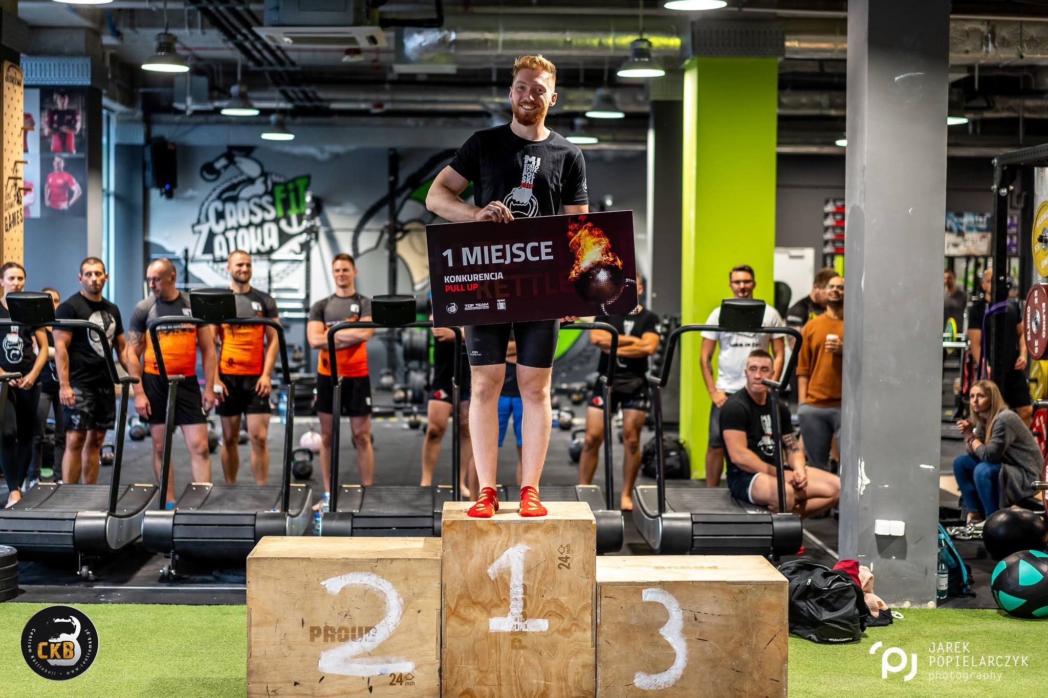 Uśmiechnięty mężczyzna z brodą, na podium z napisem „1”, trzyma tablicę z napisem „1 miejsce” w siłowni Crossfit, w tle grupa osób i bieżnie.