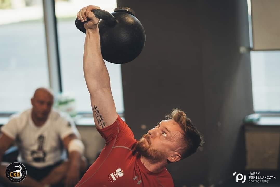 Mężczyzna w czerwonej koszulce unosi kettlebell nad głową, prezentując siłę i determinację. W tle drugi mężczyzna obserwuje trening.