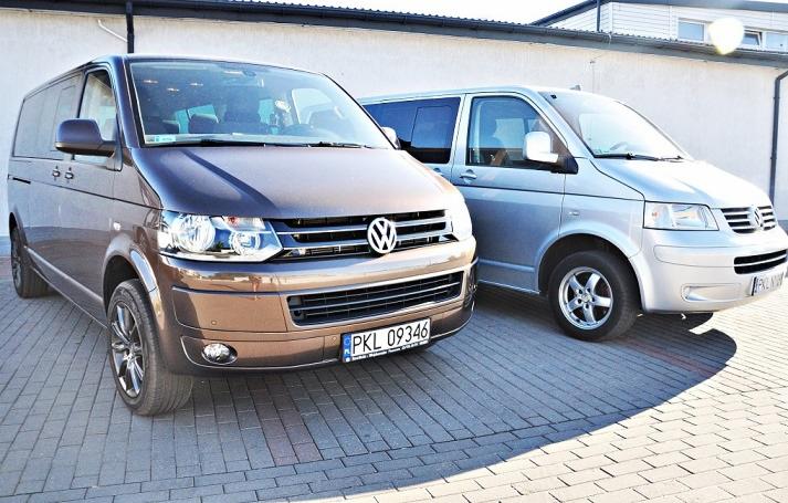 Dwa busy Volkswagen Transporter T5, jeden brązowy z przodu i srebrny w tle, zaparkowane na kostce brukowej przed białym budynkiem.