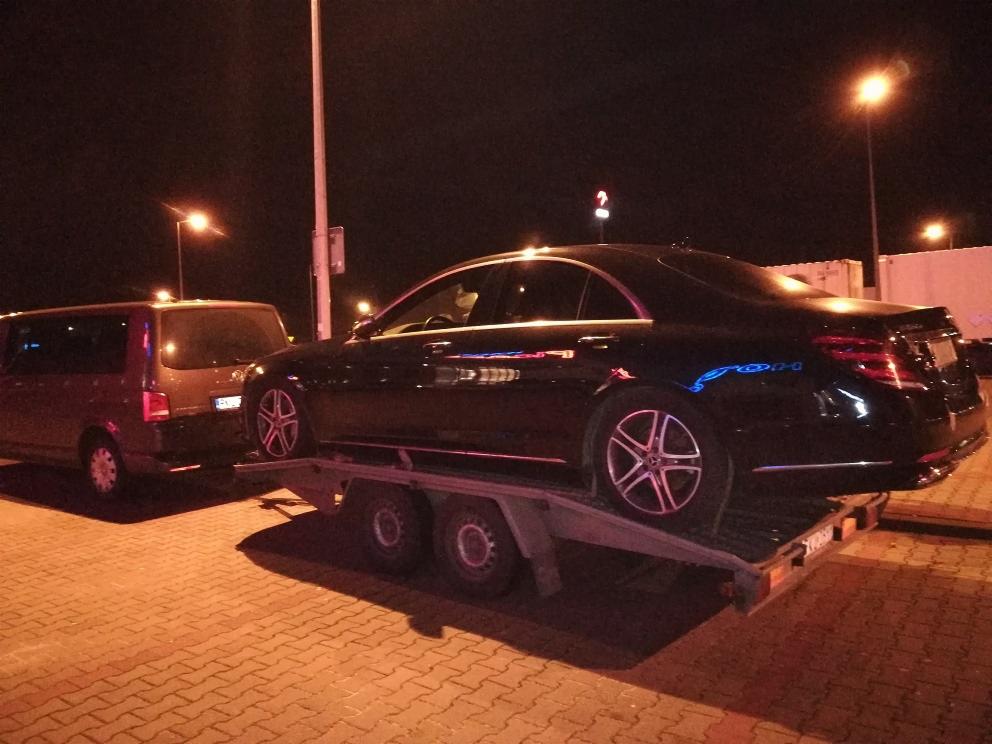 Czarny samochód osobowy na niskopodwoziowej przyczepie, holowany przez brązowy van na tle nocnego parkingu z oświetleniem.