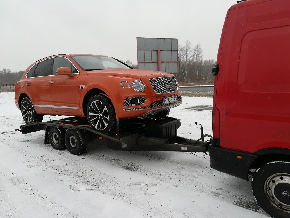 Pomarańczowy samochód osobowy marki Bentley na niskopodwoziowej przyczepie, holowanej przez czerwony samochód dostawczy. Zimowy krajobraz w tle.
