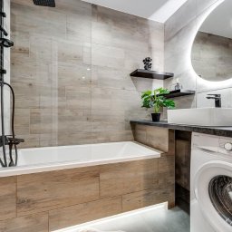 Housers sp. z o.o. - Nowoczesna łazienka z wanną obudowaną drewnopodobnymi płytkami, umywalką nablatową i pralką. Na ścianie podświetlane lustro i półki dekoracyjne.