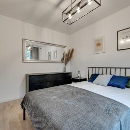 Housers sp. z o.o. - Nowoczesna sypialnia z czarnymi meblami, metalową ramą łóżka i dużym lustrem w srebrnej ramie, oświetlona lampą sufitową z żarówkami Edison.