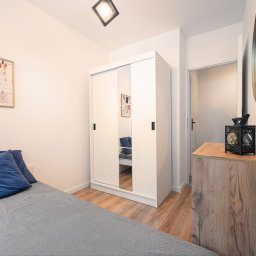 Housers sp. z o.o. - Jasne wnętrze sypialni z łóżkiem, szafą z lustrami i drewnianą komodą. Na ścianie okrągłe lustro i ramka z zdjęciami. Drewniana podłoga.