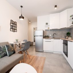 Housers sp. z o.o. - Jasne wnętrze mieszkania z aneksem kuchennym, szarą sofą i okrągłym stołem. Na ścianie wiszą dekoracyjne ramki. Widoczne białe meble kuchenne i sprzęt AGD.