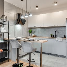 Housers sp. z o.o. - Nowoczesna, jasna kuchnia z wyspą i hokerami. Minimalistyczne szafki, szary backsplash, srebrna lodówka. Na wyspie zastawa stołowa i dekoracje. Stylowe oświetlenie.