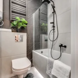 Nowoczesna łazienka z czarną armaturą, marmurowymi płytkami i puszystym dywanikiem. Uwagę zwraca podświetlenie podłogi i designerska roślina na tle grzejnika.