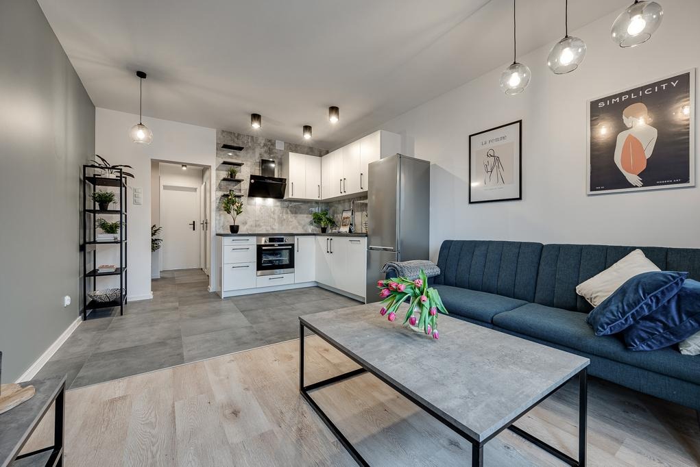 Jasne, nowocześnie urządzone wnętrze mieszkania z aneksem kuchennym, szarą sofą i betonowym stolikiem kawowym z różowymi tulipanami.