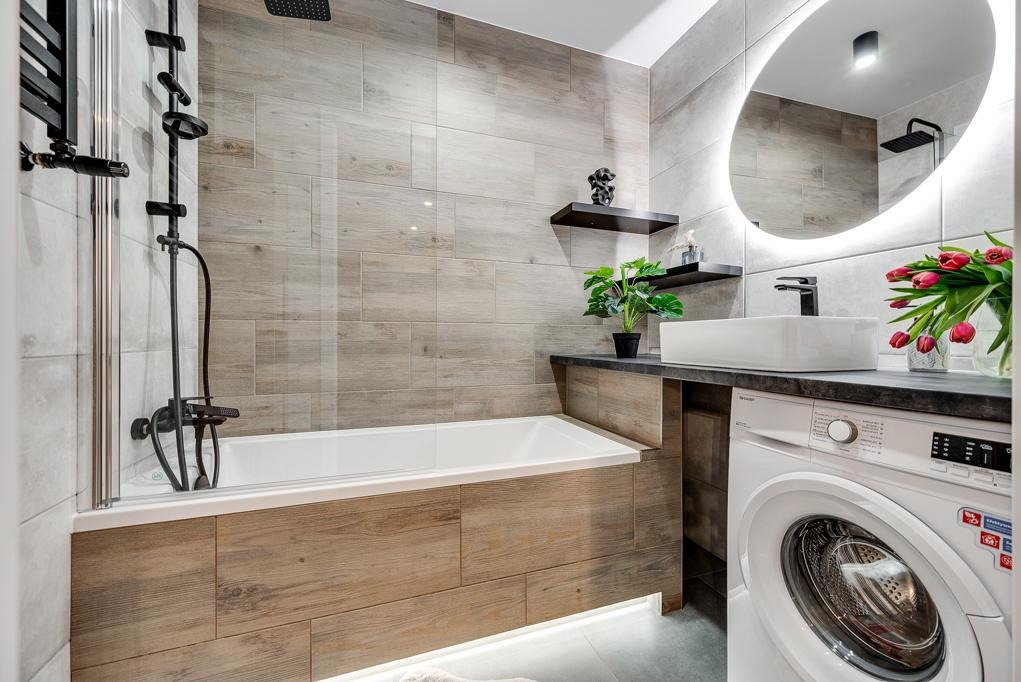 Nowoczesna łazienka z wanną obudowaną drewnopodobnymi płytkami, umywalką nablatową i pralką. Na ścianie podświetlane lustro i półki dekoracyjne.