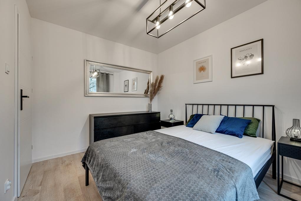 Nowoczesna sypialnia z czarnymi meblami, metalową ramą łóżka i dużym lustrem w srebrnej ramie, oświetlona lampą sufitową z żarówkami Edison.