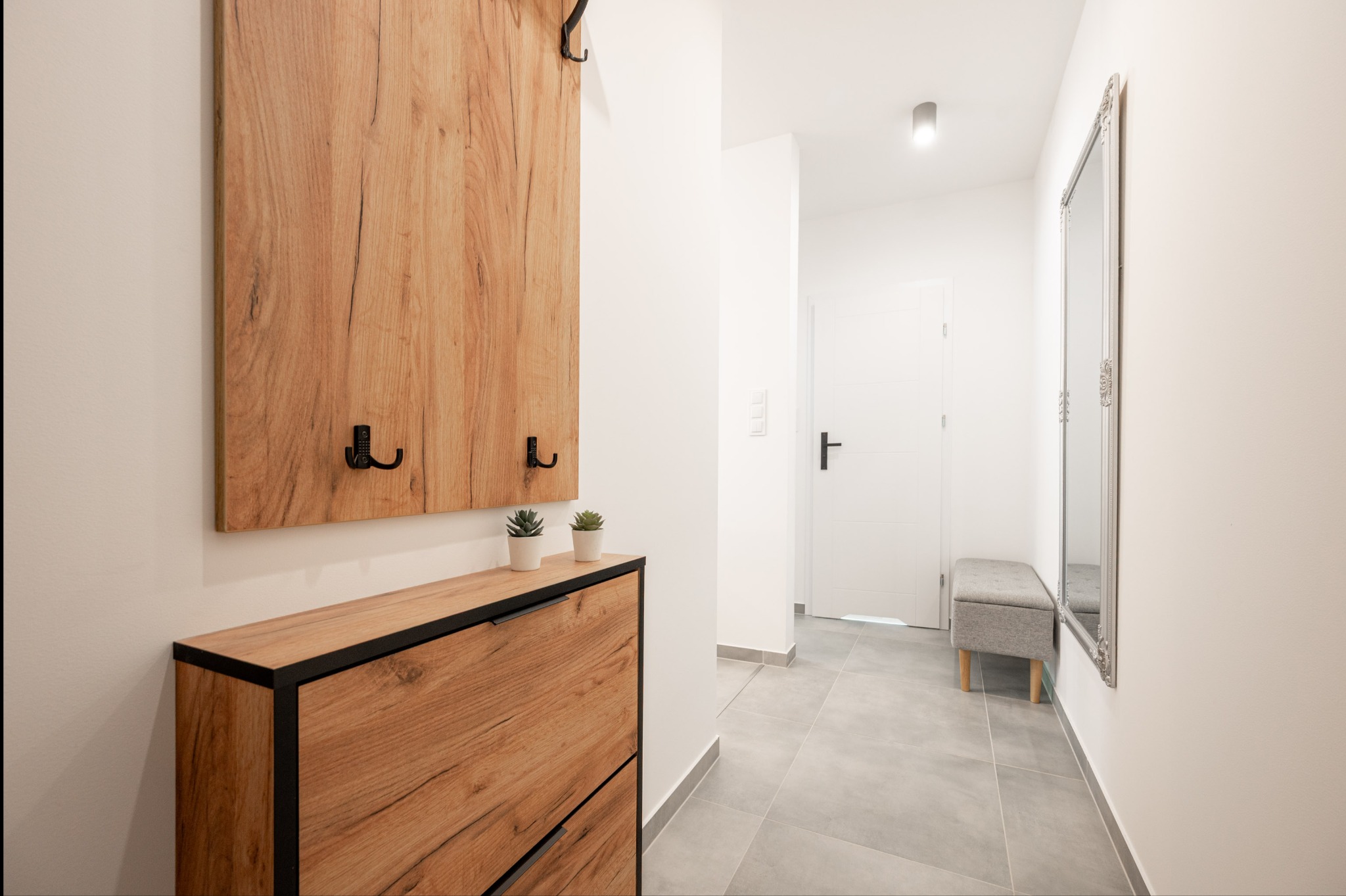 Jasne wnętrze przedpokoju z drewnianą szafką na buty, wieszakiem i lustrem w ozdobnej ramie. Minimalistyczny design, szare płytki podłogowe, białe ściany.