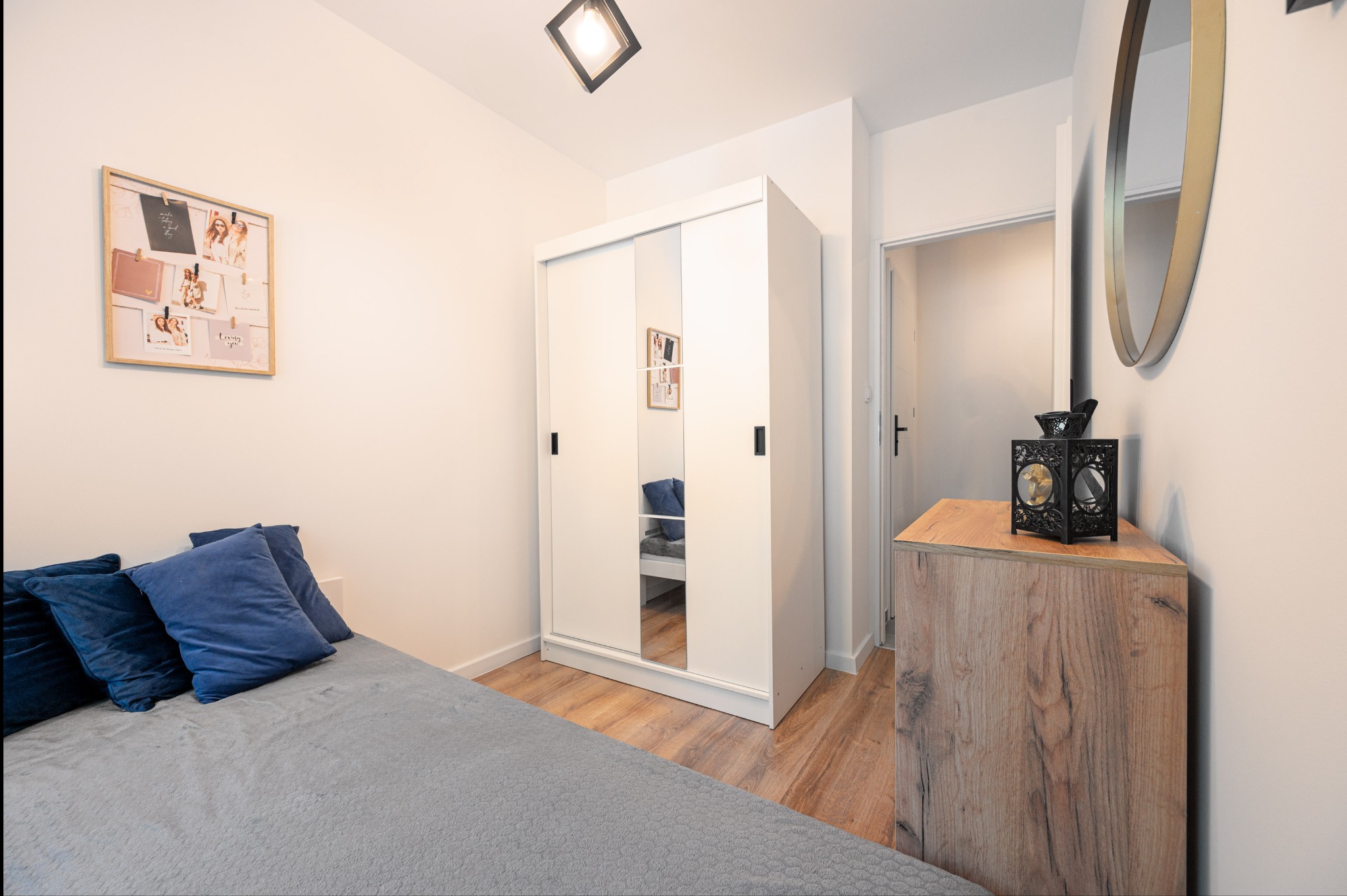 Jasne wnętrze sypialni z łóżkiem, szafą z lustrami i drewnianą komodą. Na ścianie okrągłe lustro i ramka z zdjęciami. Drewniana podłoga.