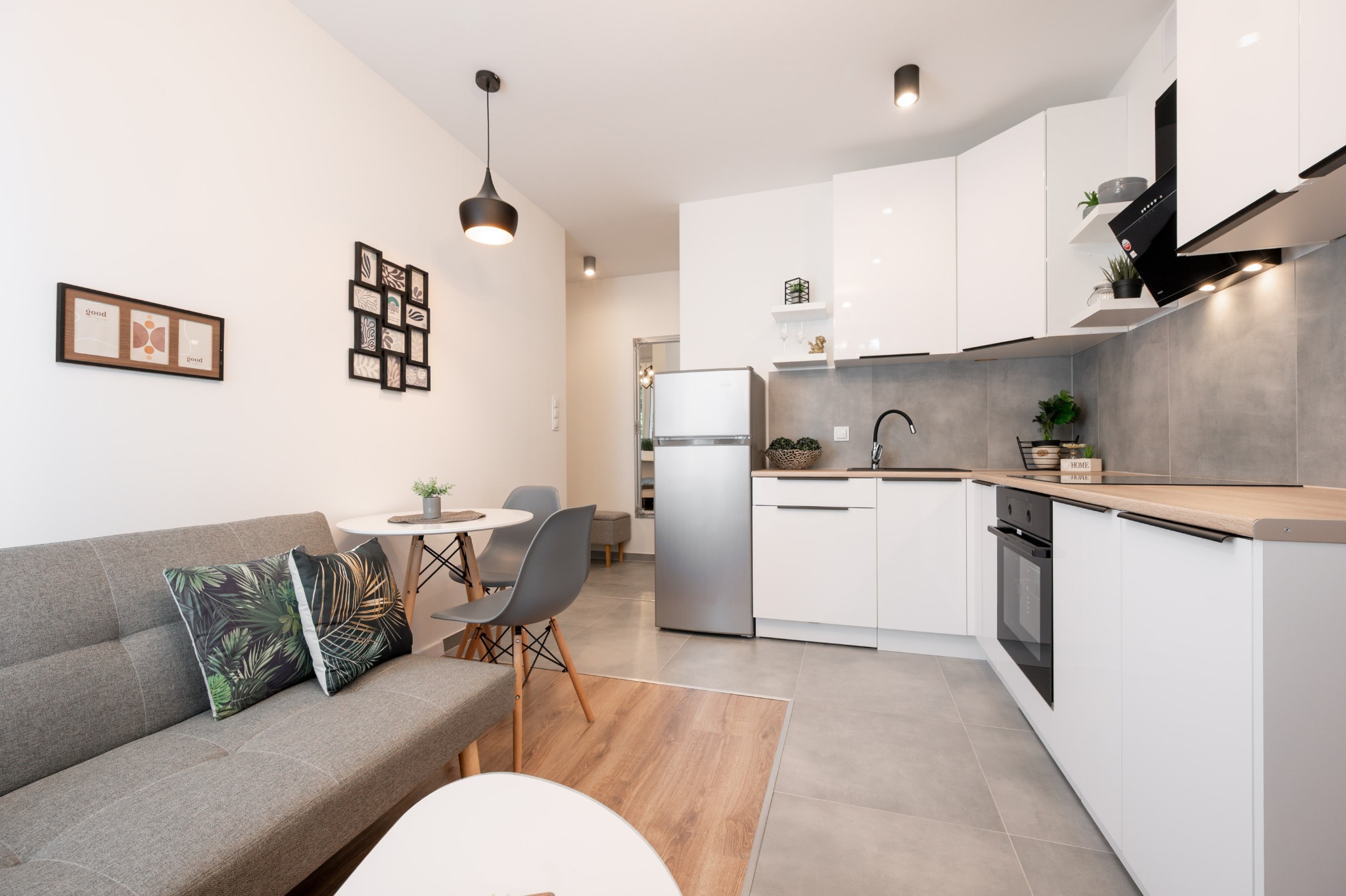 Jasne wnętrze mieszkania z aneksem kuchennym, szarą sofą i okrągłym stołem. Na ścianie wiszą dekoracyjne ramki. Widoczne białe meble kuchenne i sprzęt AGD.