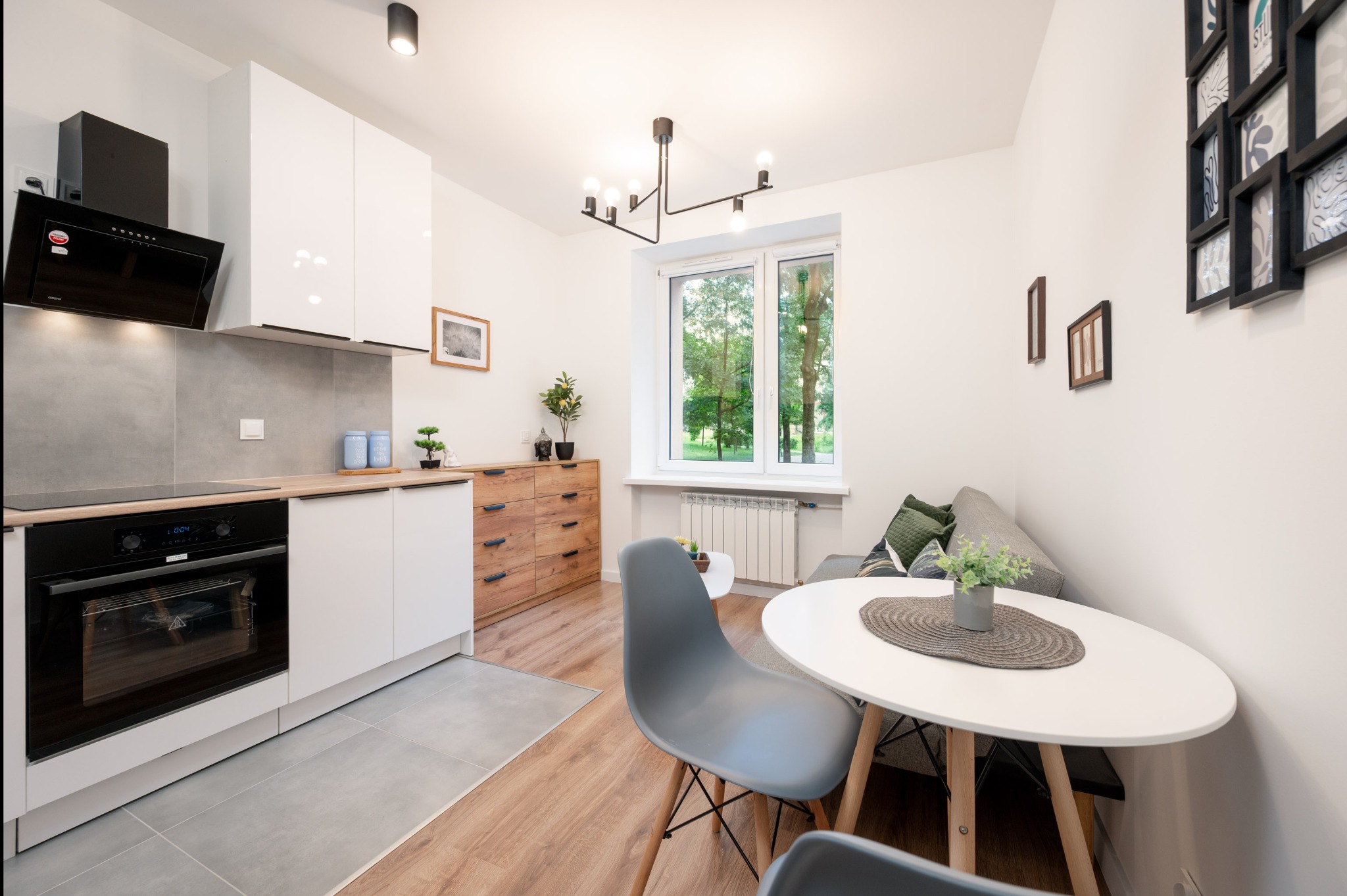 Jasne, nowocześnie urządzone wnętrze mieszkania z aneksem kuchennym, stołem i sofą. Widok na okno z zielenią. Dominuje biel i drewno.