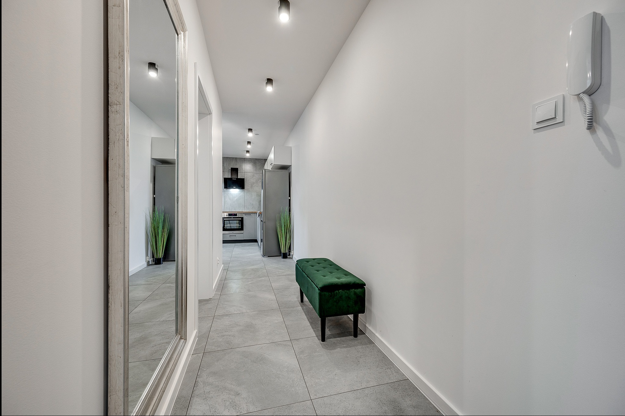 Nowoczesny korytarz z szarą podłogą, lustrem i zieloną ławką. W tle widoczna kuchnia. Minimalistyczne oświetlenie sufitowe i białe ściany.