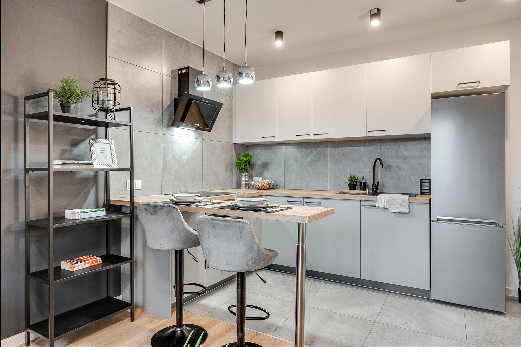 Nowoczesna, jasna kuchnia z wyspą i hokerami. Minimalistyczne szafki, szary backsplash, srebrna lodówka. Na wyspie zastawa stołowa i dekoracje. Stylowe oświetlenie.