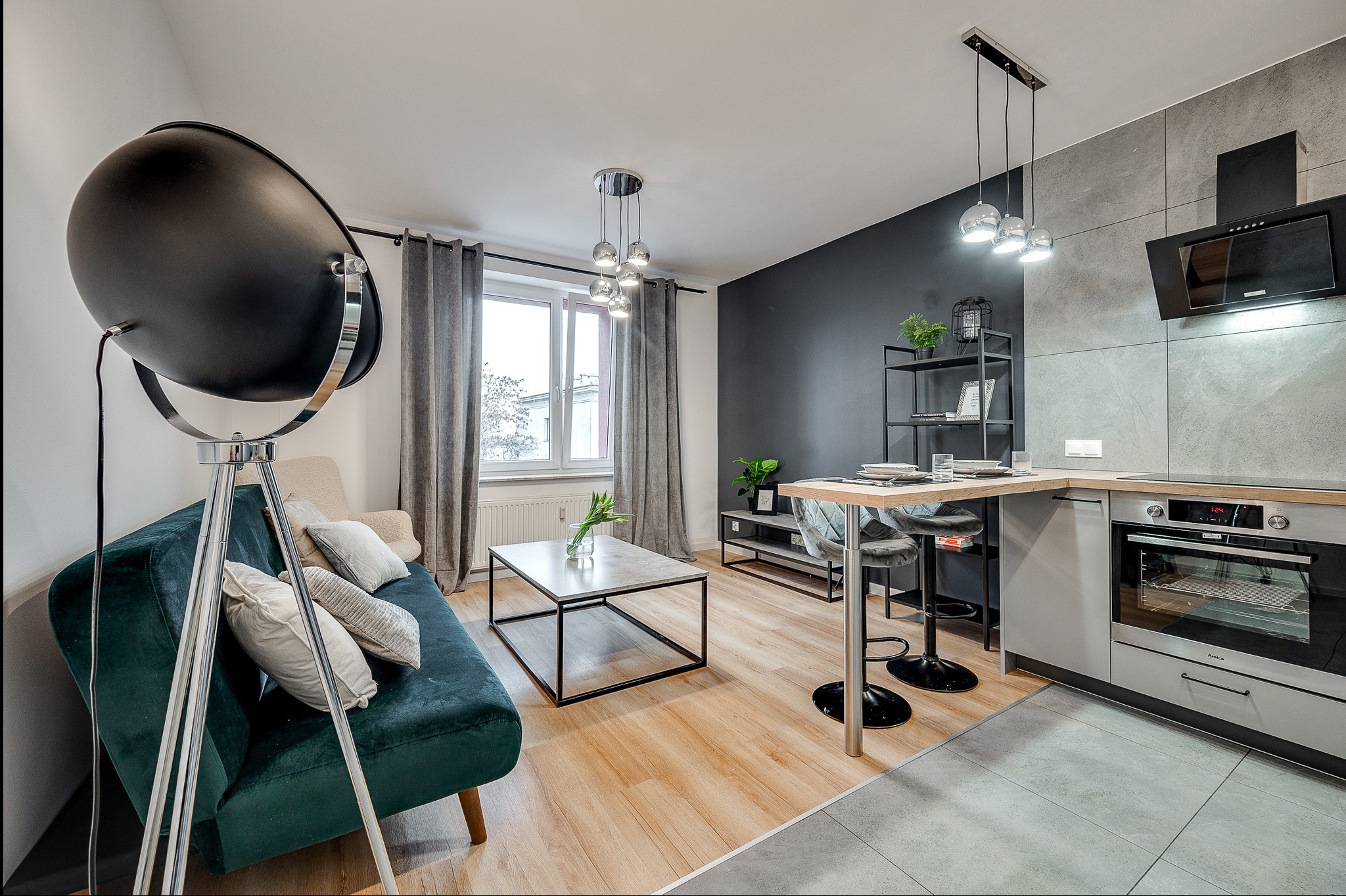 Nowoczesne wnętrze: zielona sofa, lampa reflektorowa, stolik kawowy, aneks kuchenny z barkiem. Aranżacja mieszkania z widokiem na okno i stylowe dodatki.