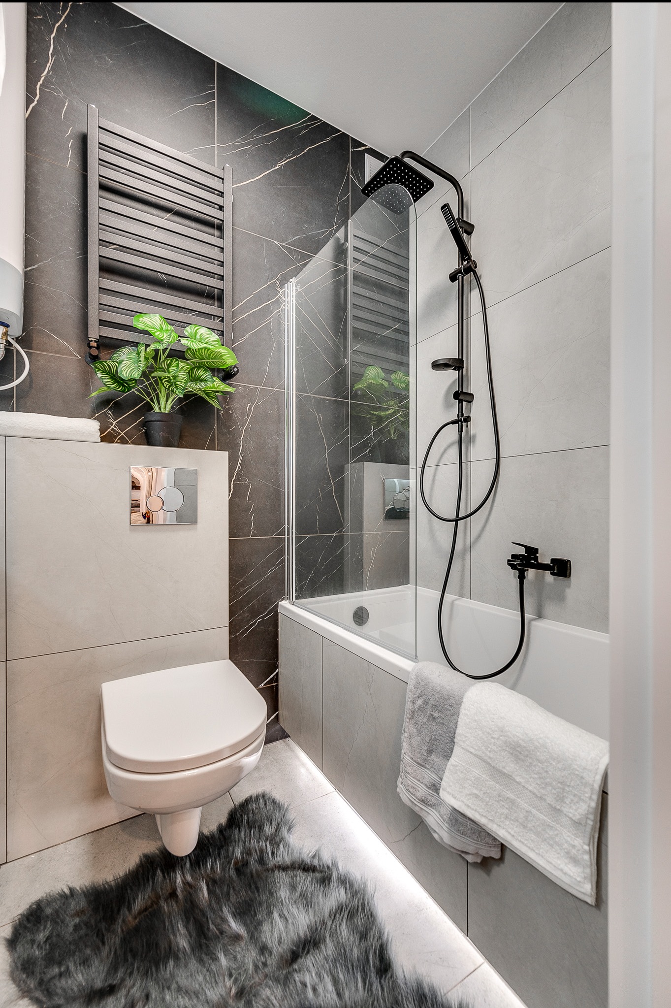 Nowoczesna łazienka z czarną armaturą, marmurowymi płytkami i puszystym dywanikiem. Uwagę zwraca podświetlenie podłogi i designerska roślina na tle grzejnika.