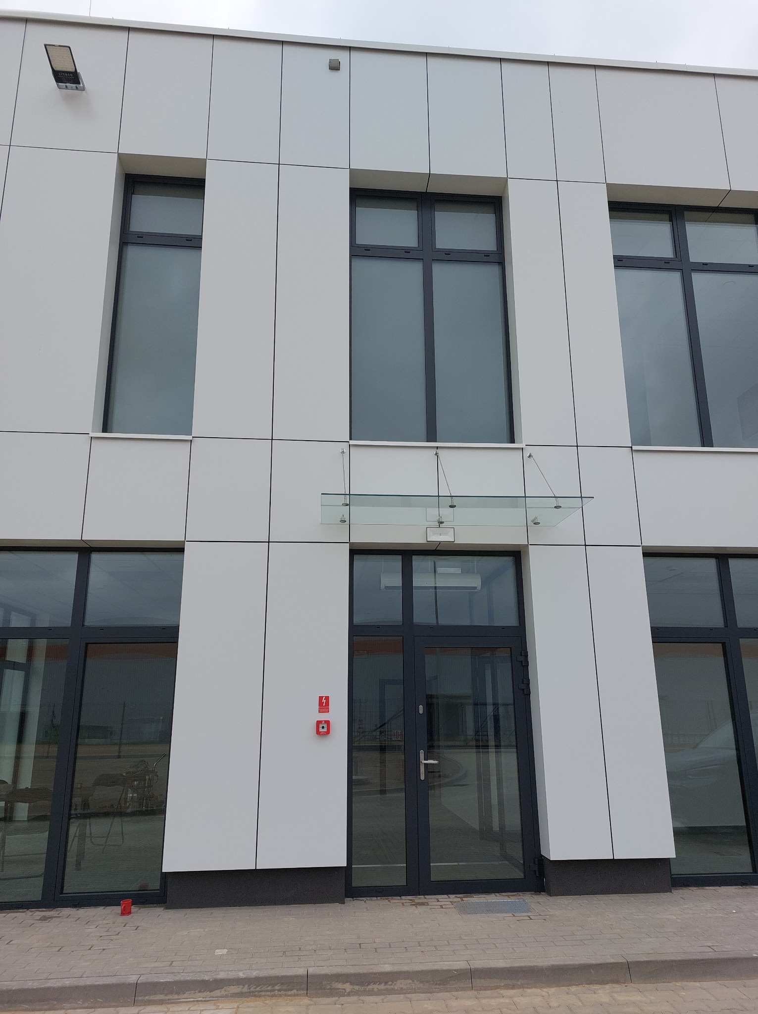 Elewacja biurowca z szarymi oknami i drzwiami, wykończona panelami. Nad wejściem szklany daszek. Widoczne detale jak lampa i czujnik przeciwpożarowy.