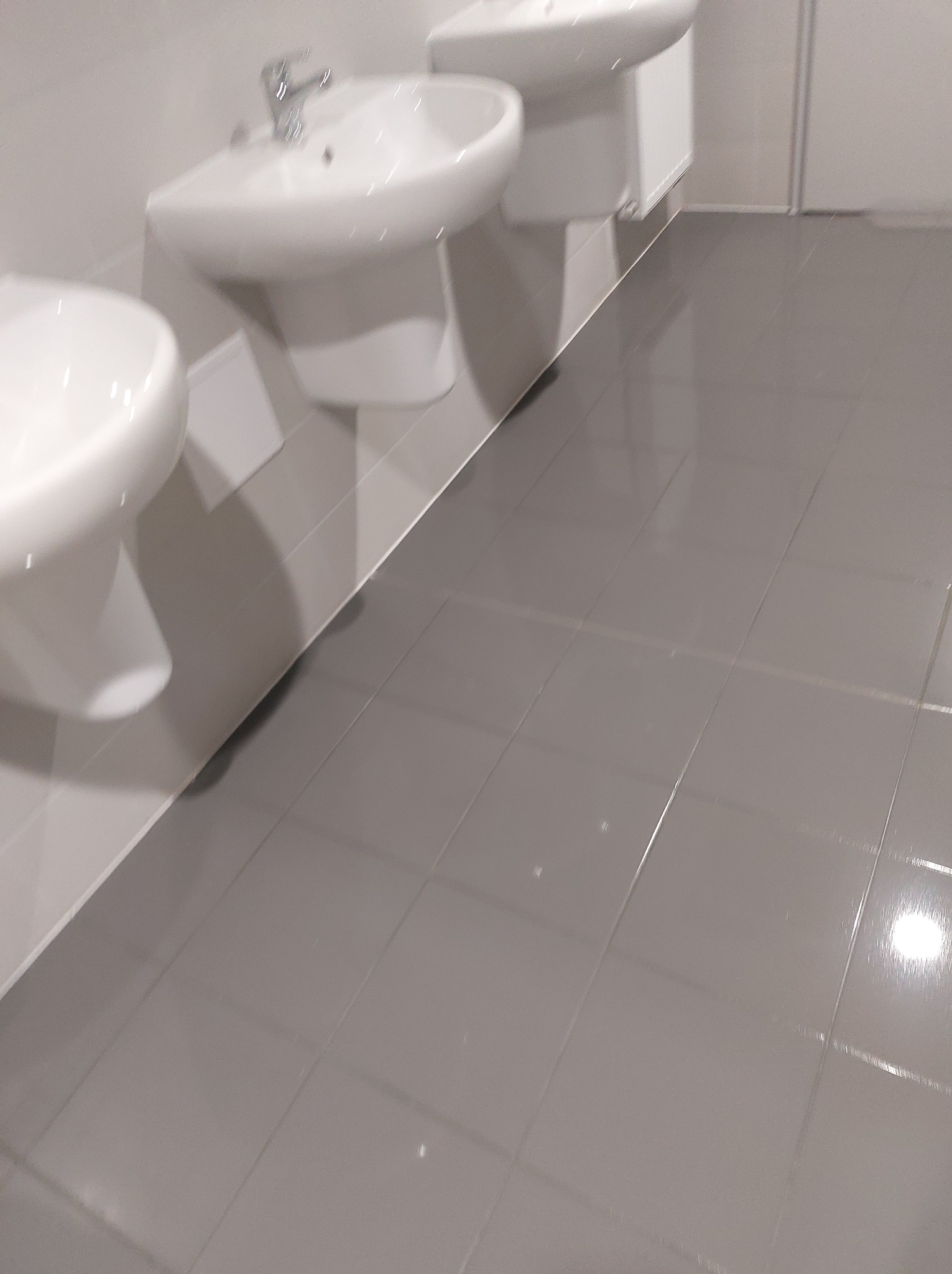 Błyszcząca, szara podłoga z płytek w łazience z trzema umywalkami. Widok z niskiej perspektywy, podkreślający czystość i połysk powierzchni.