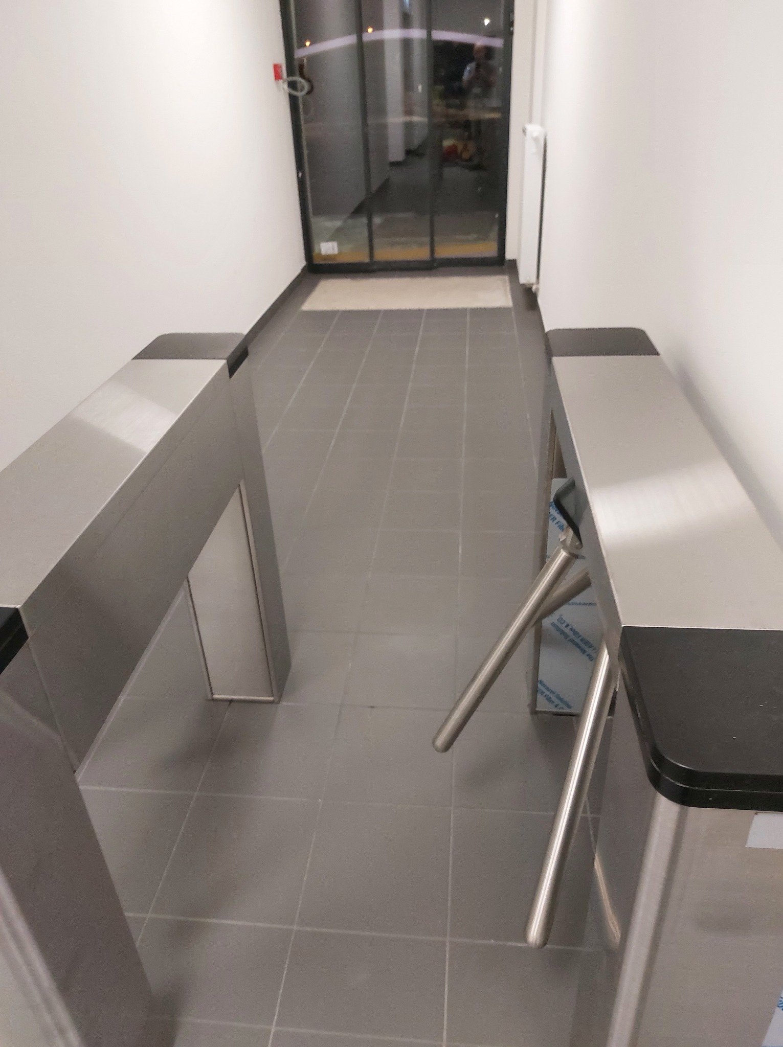 Szare płytki podłogowe w biurowcu z widokiem na szklane drzwi i nowoczesny, metalowy kołowrót wejściowy z czarnymi elementami wykończeniowymi.