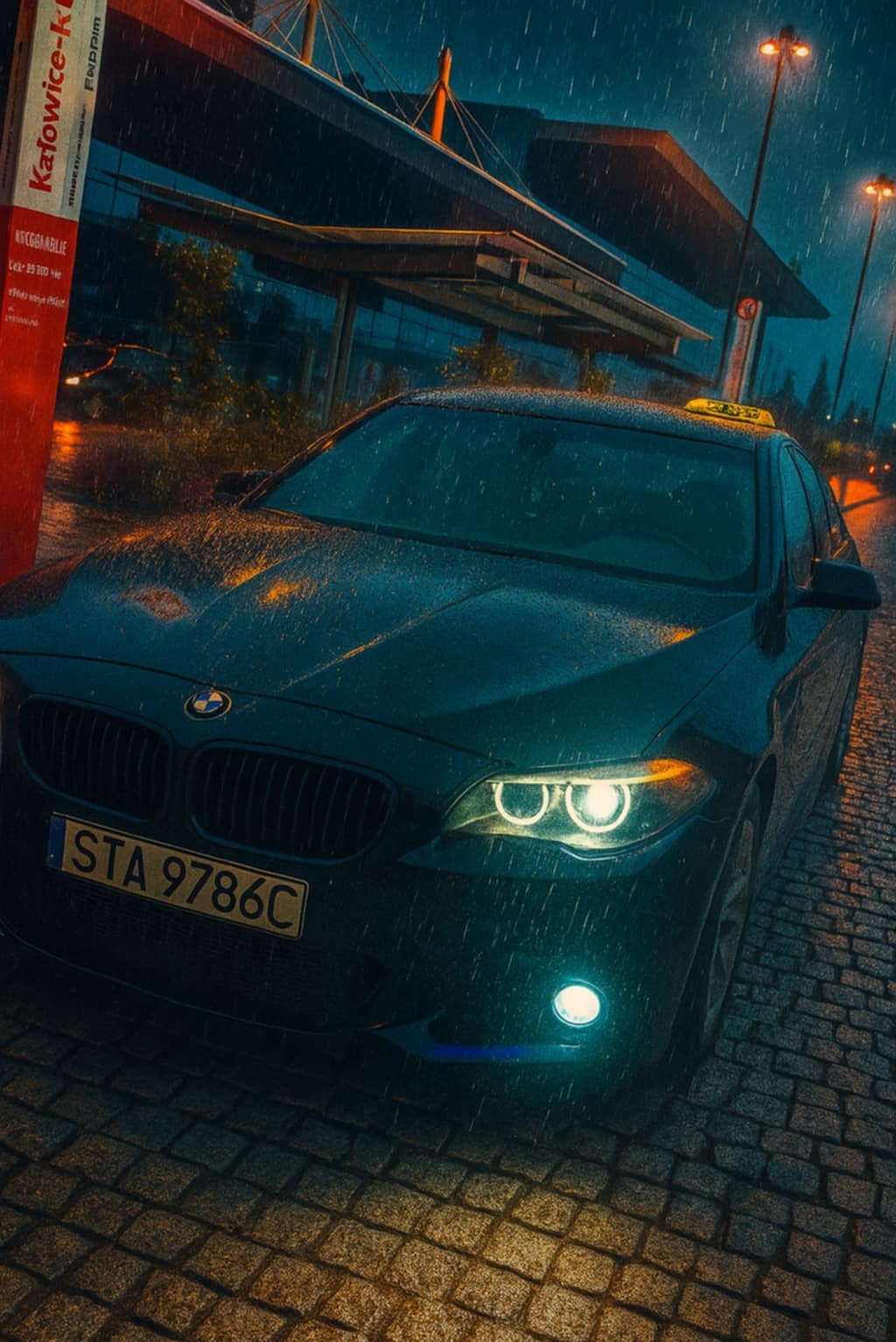 Czarna taksówka BMW, oświetlona deszczem, stoi na brukowanej ulicy w Katowicach pod przystankiem autobusowym nocą. Widoczne krople deszczu i światła uliczne.