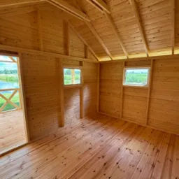 Wnętrze nowej, drewnianej konstrukcji z widokiem na jezioro przez otwarte drzwi i dwa okna. Ściany, podłoga i sufit wykonane z jasnego drewna.