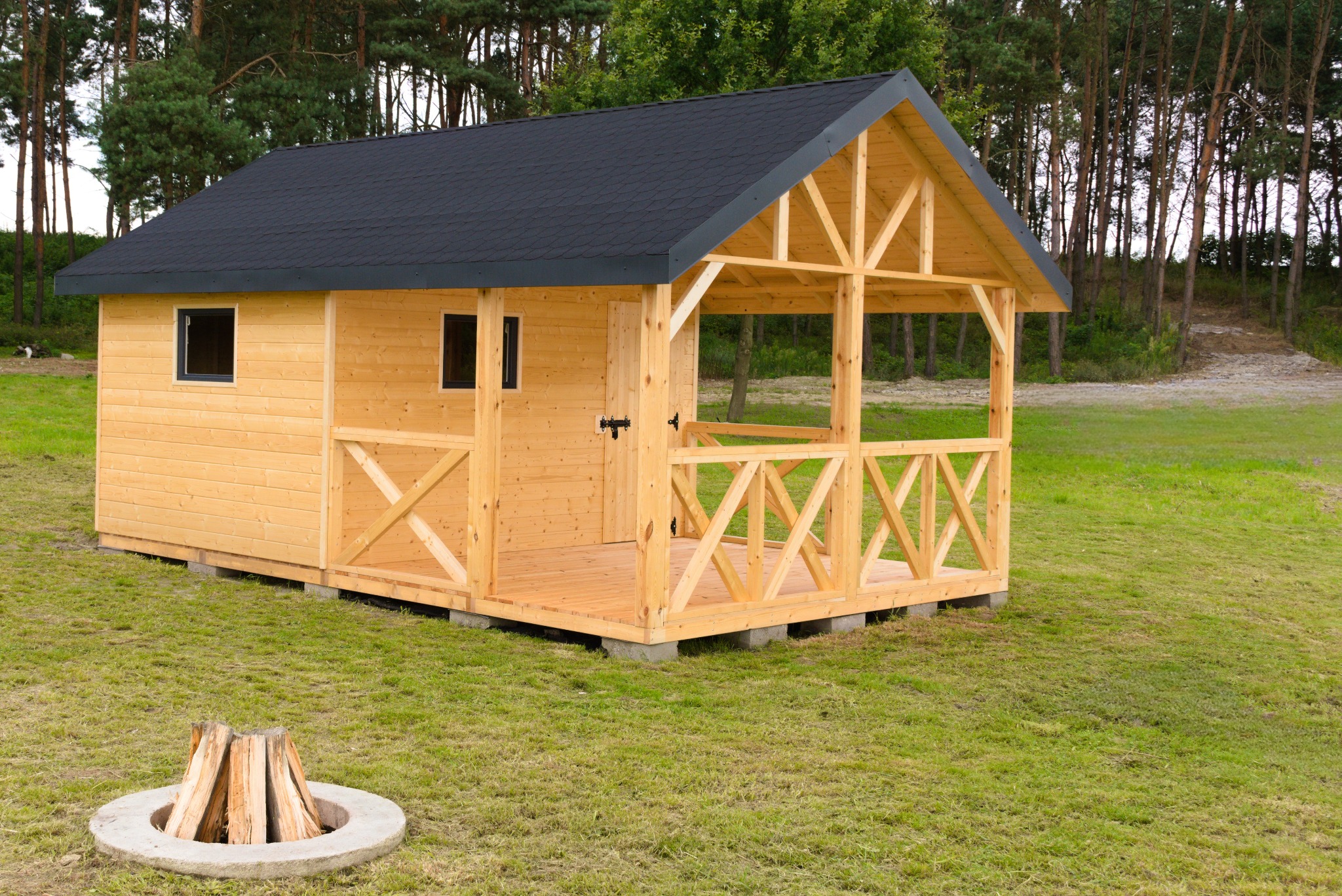 Nowoczesna, drewniana altana z zadaszeniem i tarasem, usytuowana na trawniku obok betonowego paleniska. Widoczne detale konstrukcyjne i czarne pokrycie dachu.