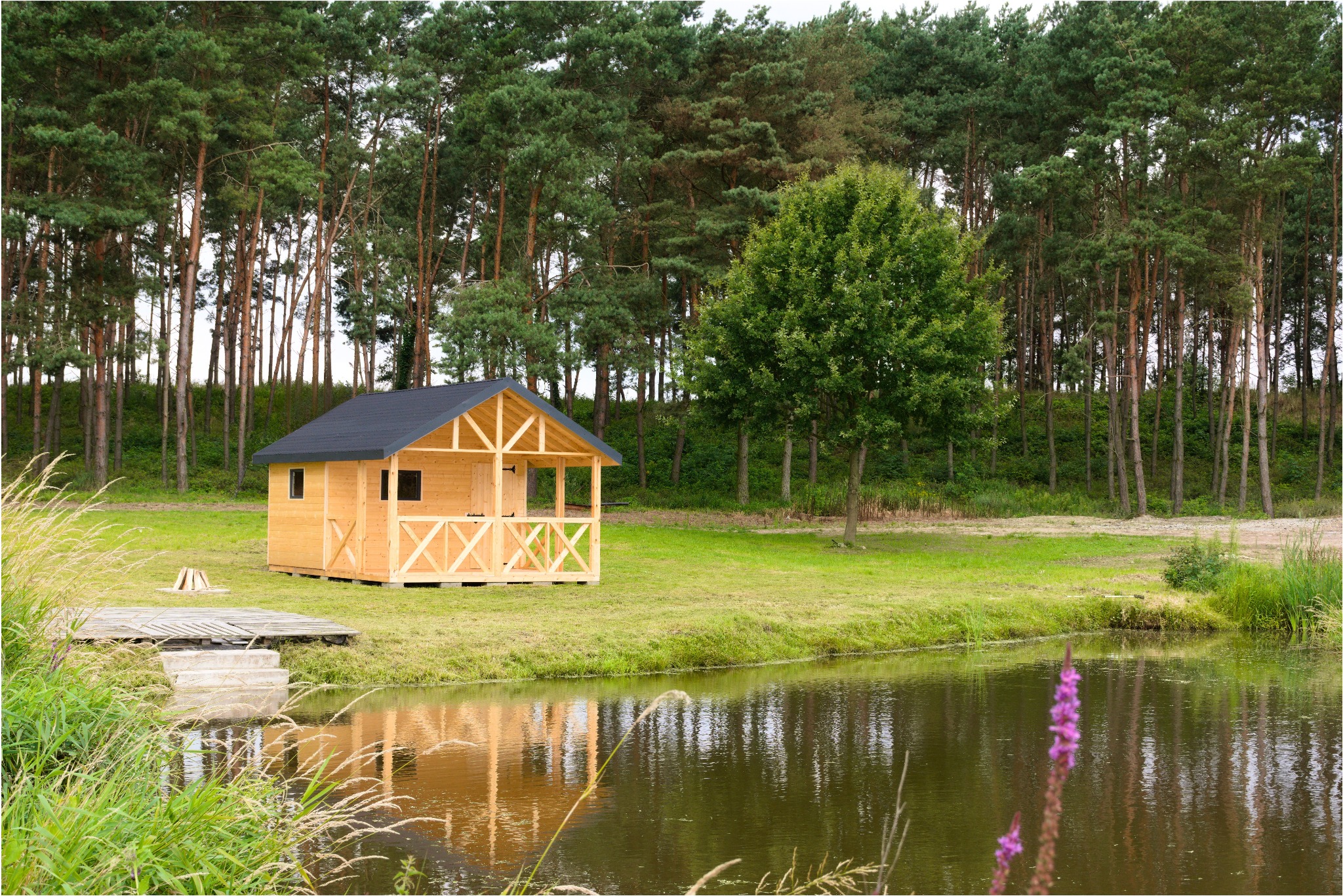 Drewniana altana z czarnym dachem nad brzegiem stawu, otoczona lasem. Pomost z zejściem do wody. Naturalne otoczenie, trawa i drzewa odbijające się w tafli wody.