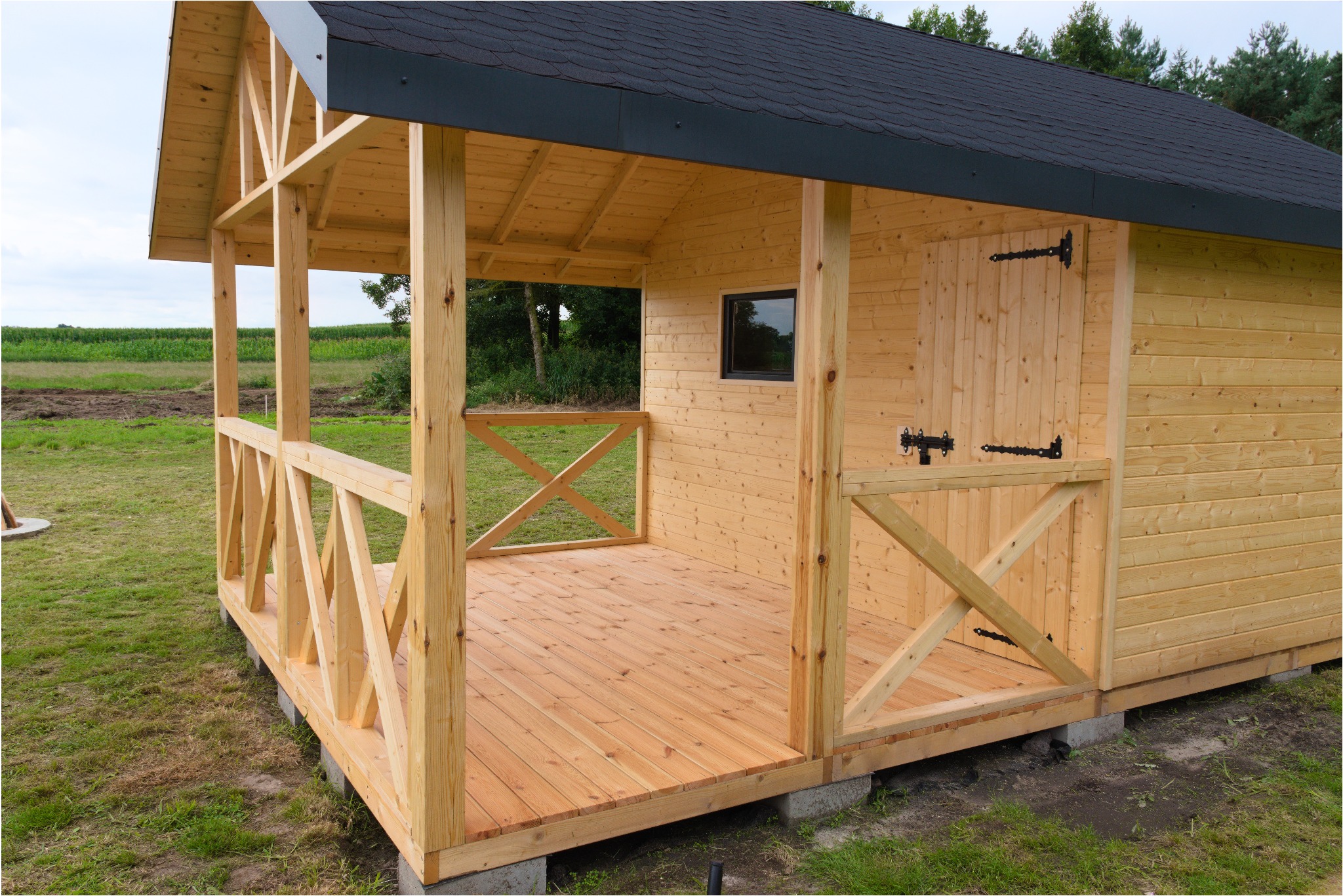 Drewniana altana z czarnym dachem krytym gontem bitumicznym, z podłogą z desek, na betonowych bloczkach, z dekoracyjną balustradą w kształcie litery X.