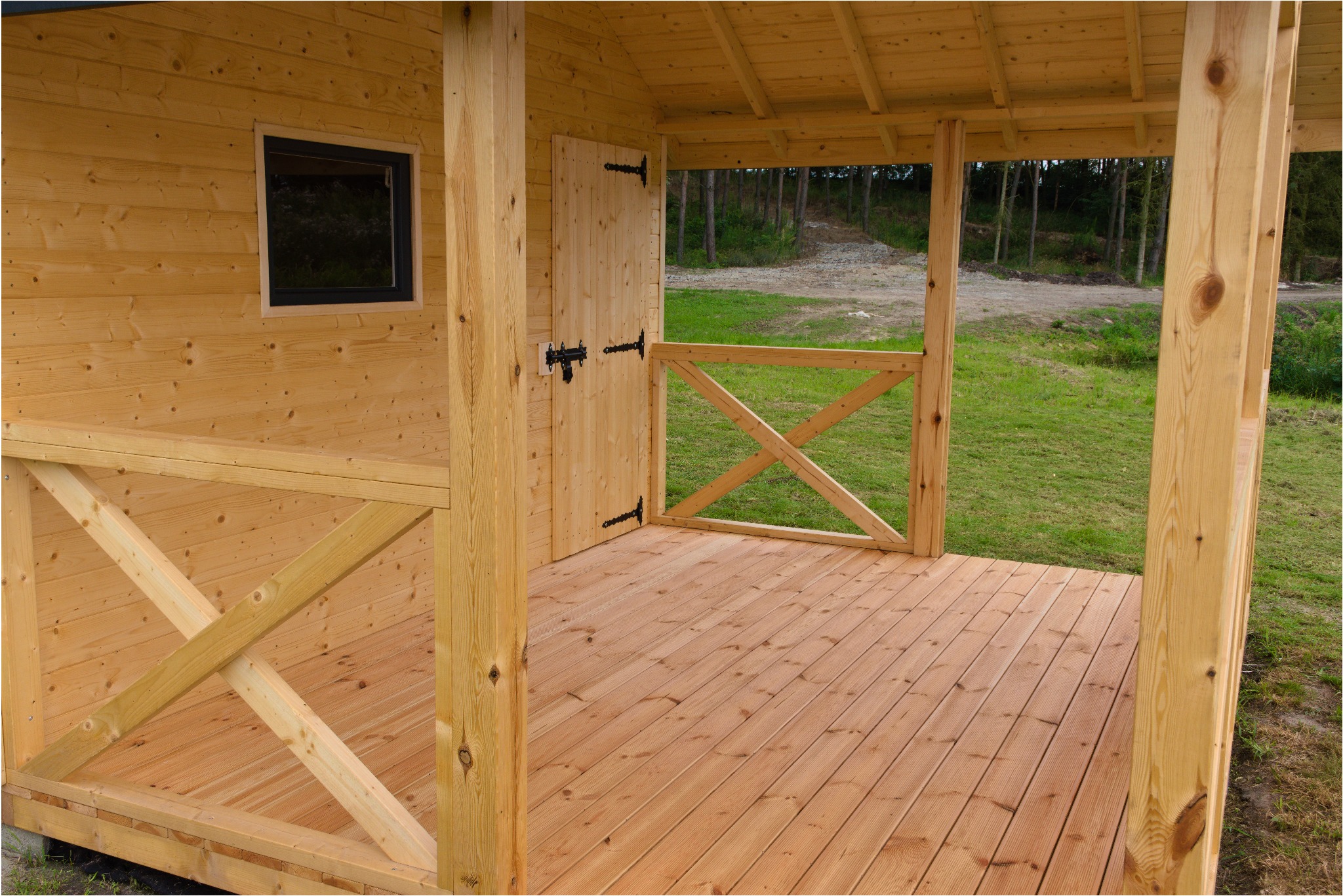 Drewniana altana z podłogą z desek, otwartymi drzwiami z czarnymi zawiasami i widokiem na zieleń. Naturalne drewno, prosta konstrukcja, światło dzienne.