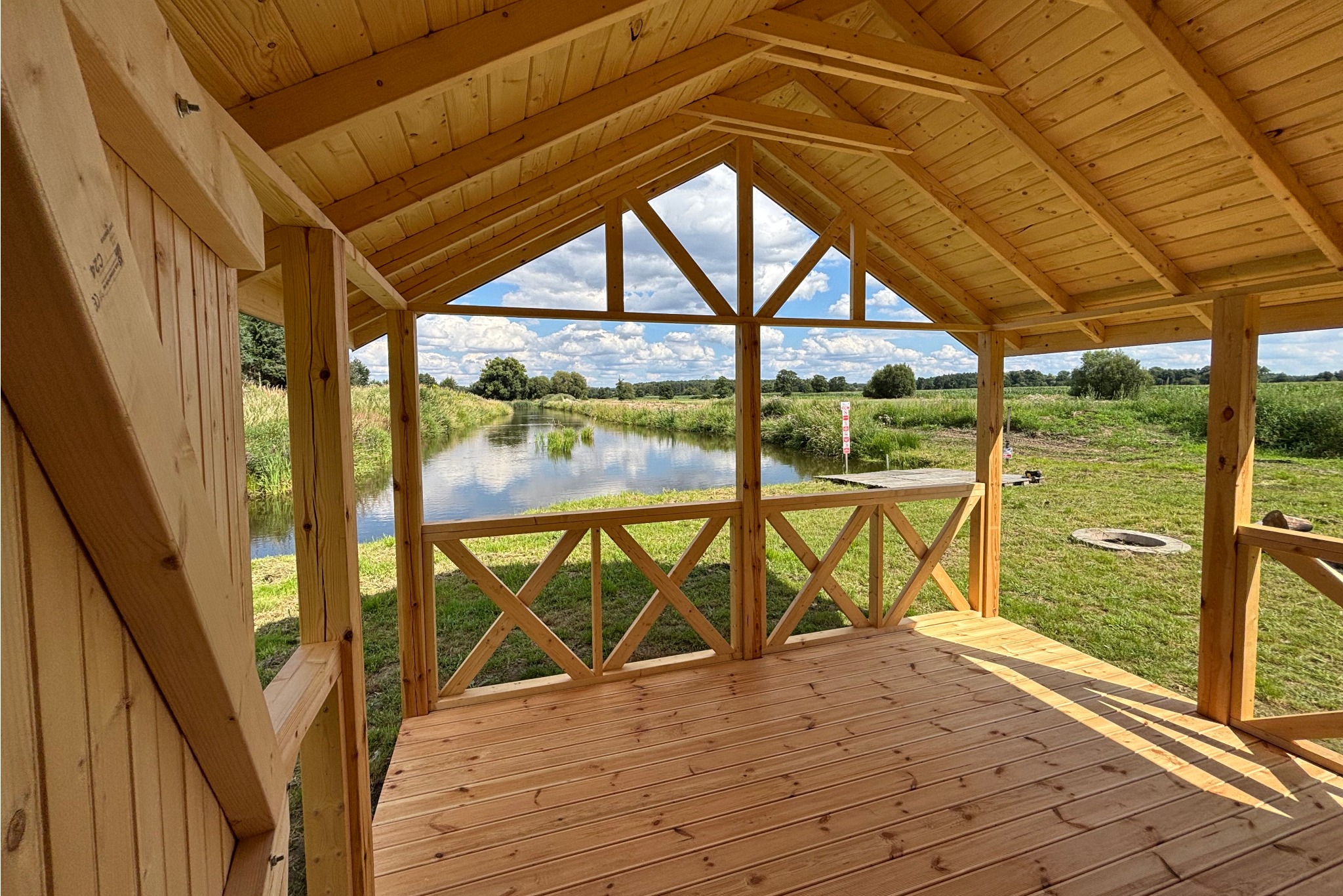 Drewniana altana z widokiem na rzekę. Konstrukcja z jasnego drewna, z ażurowym dachem i balustradą. W tle zielona łąka i woda odbijająca niebo.