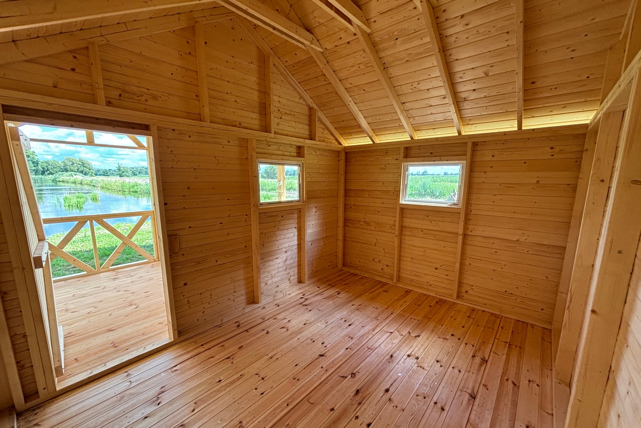 Wnętrze nowej, drewnianej konstrukcji z widokiem na jezioro przez otwarte drzwi i dwa okna. Ściany, podłoga i sufit wykonane z jasnego drewna.