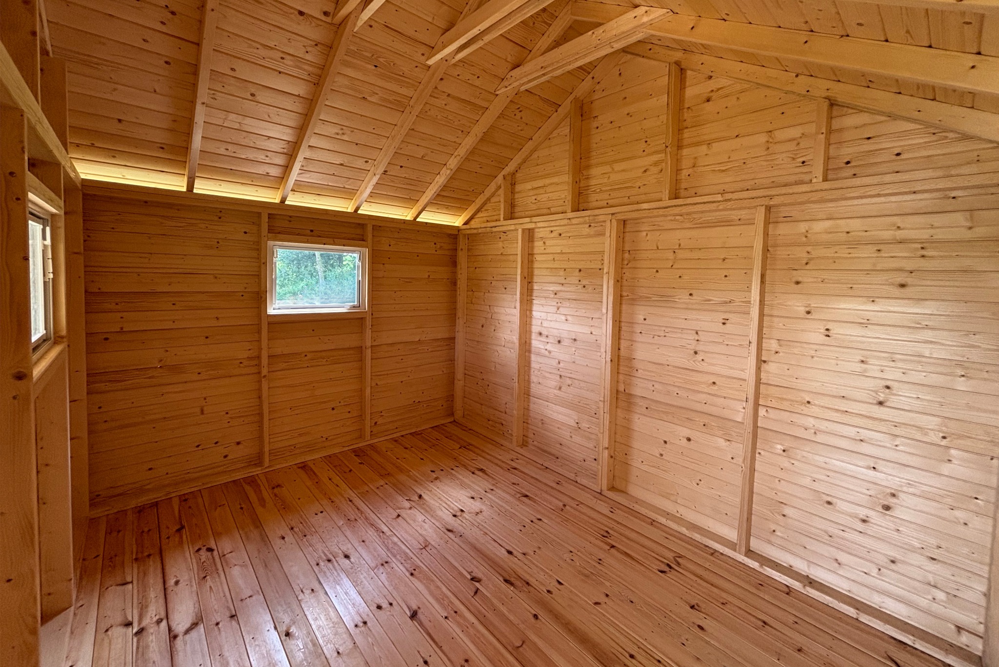 Wnętrze drewnianej konstrukcji z oknami, ukazujące surowe deski ścian, podłogi i skośnego dachu. Naturalne światło wpada przez okna, podkreślając teksturę drewna.