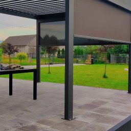 K2-ROL KAROL KRUSZCZAK - Nowoczesna pergola z rozwijaną przesłoną w kolorze grafitowym, na tle zielonego ogrodu i budynku gospodarczego. Widoczne elementy konstrukcyjne i wykończenie tarasu.