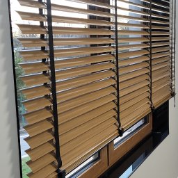 K2-ROL KAROL KRUSZCZAK - Drewniane żaluzje poziome z czarnymi sznurkami w oknie, widok z boku, z zewnątrz widoczny fragment zieleni. Naturalne światło podkreśla strukturę drewna.