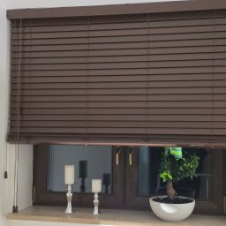 K2-ROL KAROL KRUSZCZAK - Brązowe żaluzje drewniane zamontowane w oknie, z widocznymi sznurkami do regulacji. Na parapecie dekoracje: świeczniki i bonsai. Ujęcie z bliska, wnętrze.