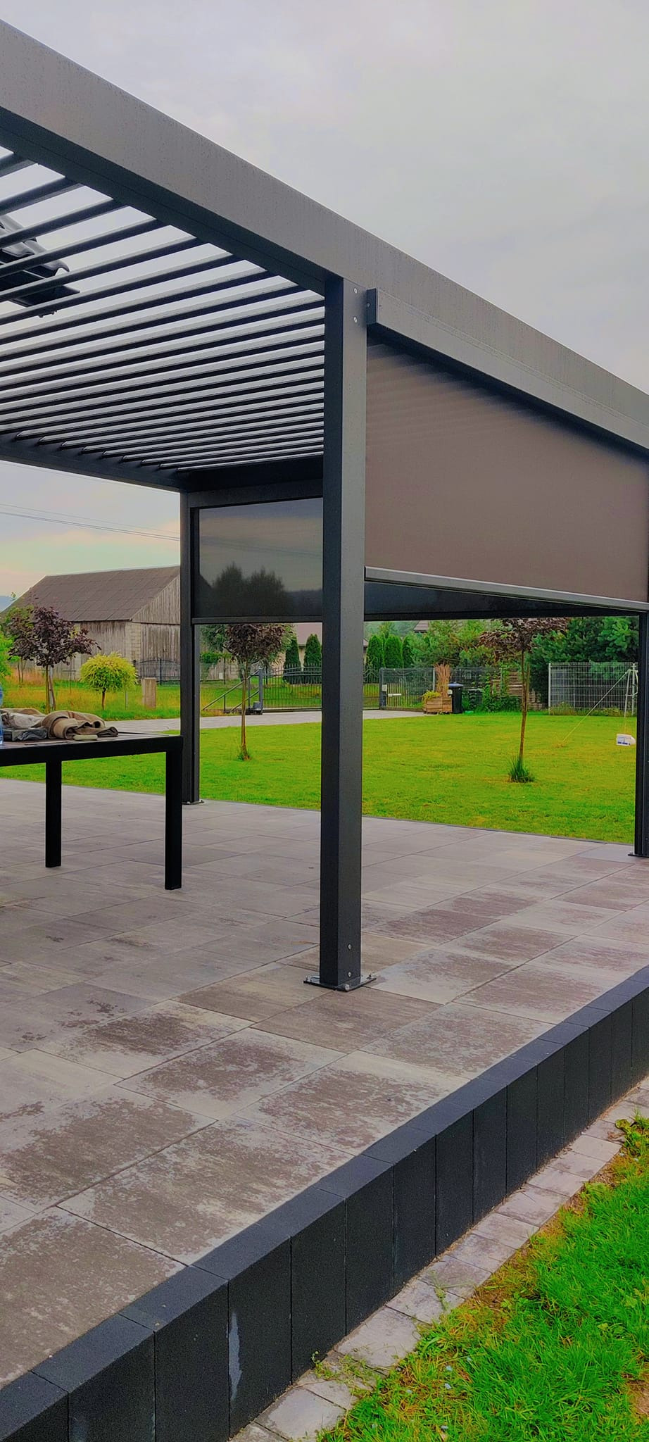 Nowoczesna pergola z rozwijaną przesłoną w kolorze grafitowym, na tle zielonego ogrodu i budynku gospodarczego. Widoczne elementy konstrukcyjne i wykończenie tarasu.