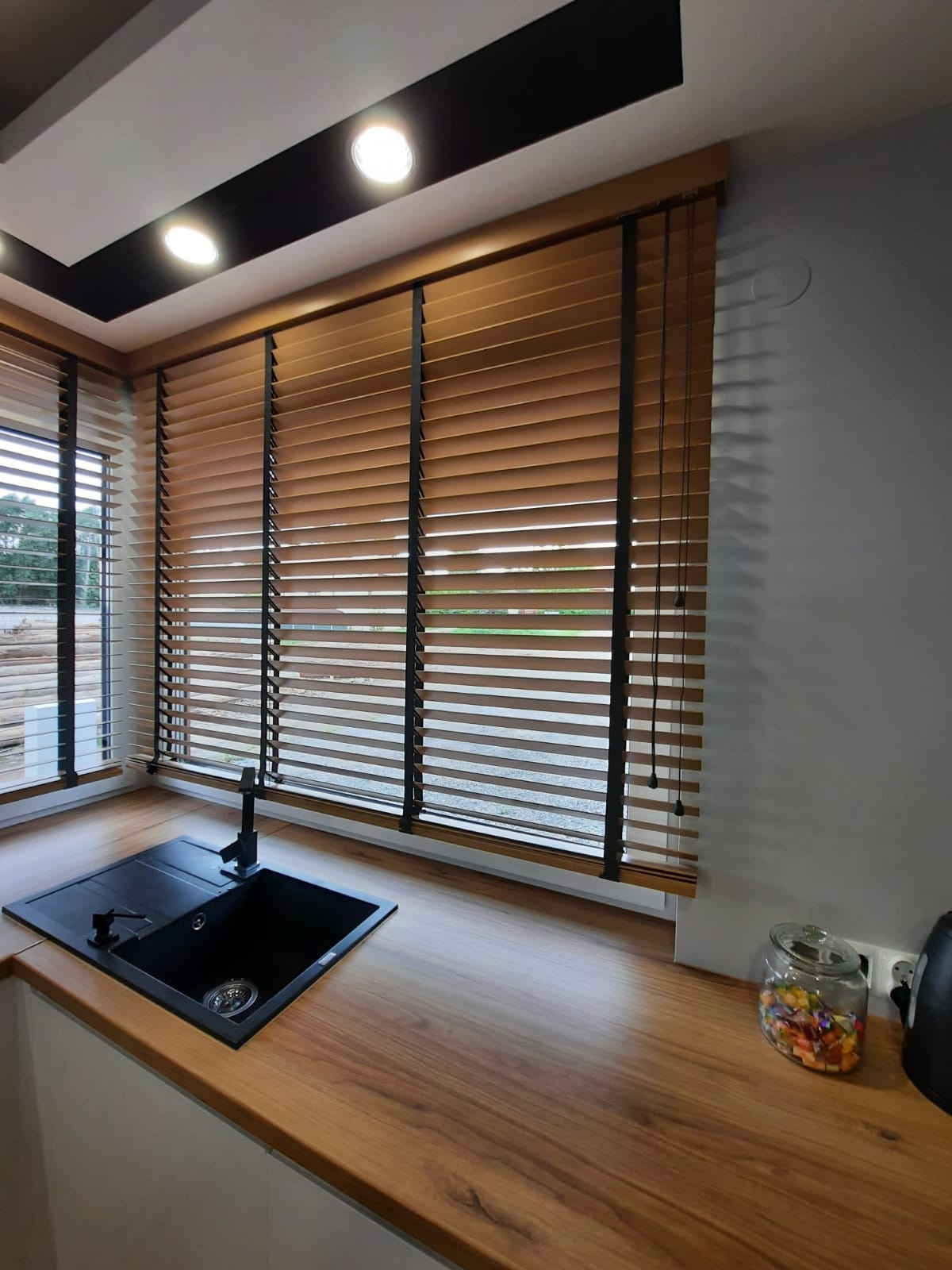 Drewniane żaluzje poziome w kuchni w Woli Kopcowej. Naturalne światło, czarne akcenty, widok na zewnątrz. Nowoczesny design, ciepłe odcienie drewna.