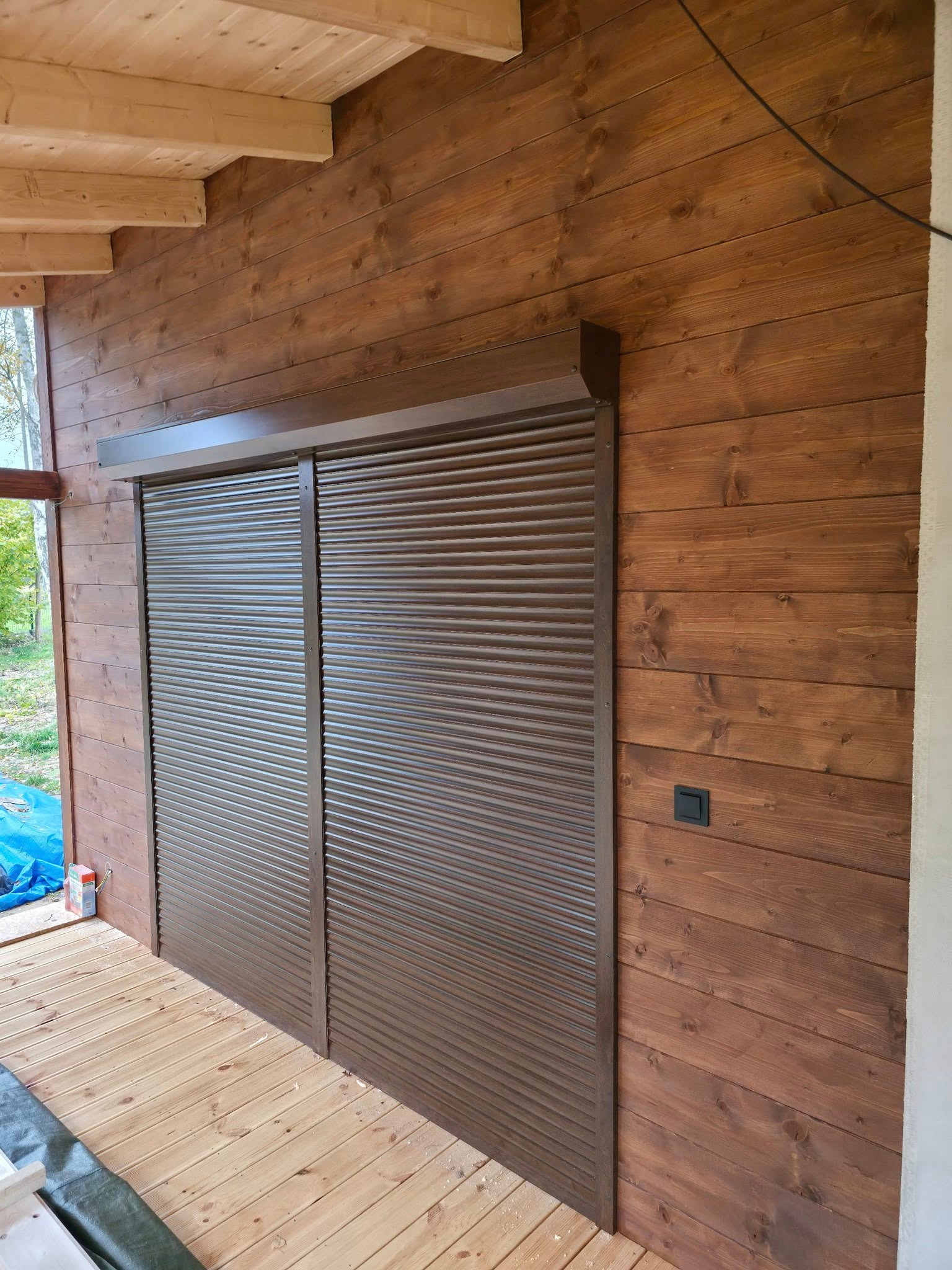 Brązowe, poziome osłony okienne na drewnianej ścianie z desek, widoczne z bliska. Na podłodze deski, w tle zieleń. Zamknięte osłony w kolorze brązu.