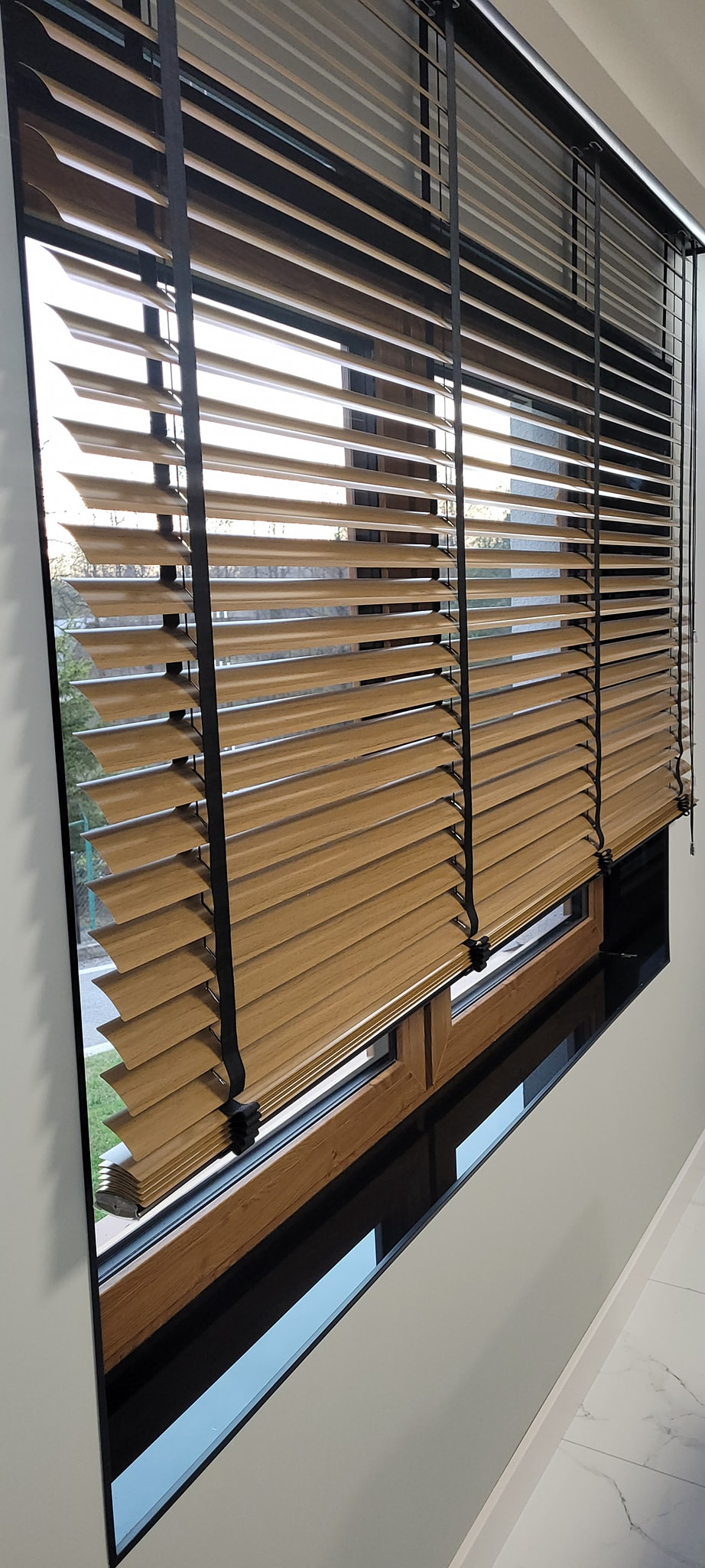 Drewniane żaluzje poziome z czarnymi sznurkami w oknie, widok z boku, z zewnątrz widoczny fragment zieleni. Naturalne światło podkreśla strukturę drewna.
